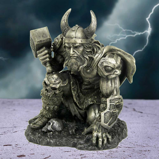 A detailed statue depicts a Viking warrior crouching, holding a hammer and wearing a horned helmet, surrounded by skulls on a stormy background with lightning.