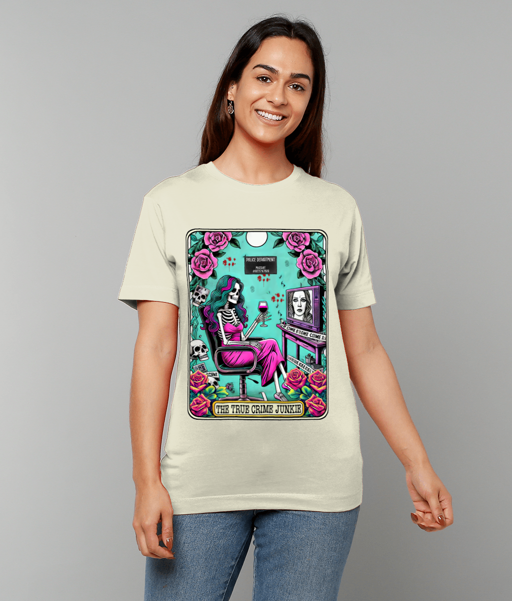 T-shirt graphic depicts a skeleton woman in a pink dress sipping wine while seated and watching a mugshot on a monitor surrounded by roses and skulls, worn by a smiling woman against a plain gray background. Text: THE TRUE CRIME JUNKIE