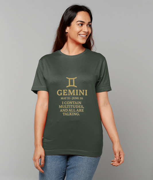Green T-shirt displaying a gold Gemini symbol and slogan worn by a smiling woman standing against a plain light-gray backdrop; text reads: "GEMINI MAY 21 - JUNE 20 I CONTAIN MULTITUDES, AND ALL ARE TALKING."