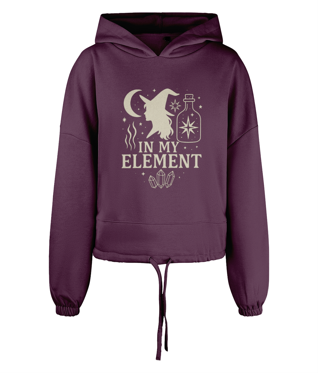 A plum cropped hoodie displays a cream witch-profile graphic with moon, potion bottle, crystals and the text "IN MY ELEMENT" centered against a plain black background.