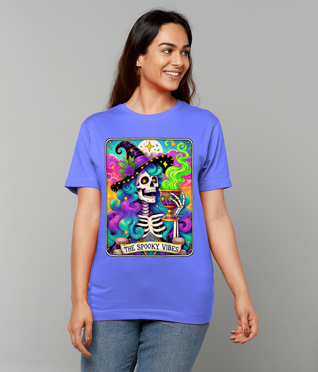 Skeleton-witch illustration on a lavender T-shirt smiles and holds a steaming goblet while colorful smoke swirls around her; wearer stands against a plain gray background; text on design reads "THE SPOOKY VIBES".