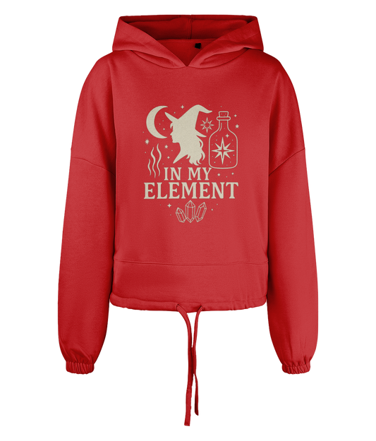 Red hoodie features a stylized witch profile, crescent moon, potion bottle, crystals and the text "IN MY ELEMENT" centered on the chest against a plain black background.