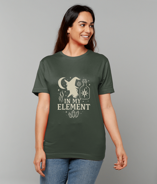 A smiling woman wears a dark green T-shirt printed with a witch silhouette, moon, potion bottle, crystals and the text IN MY ELEMENT against a plain gray background.