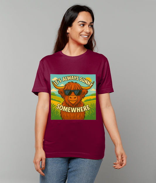 T-shirt shows a cartoon Highland cow wearing sunglasses and smiling in a sunny field, worn by a smiling woman standing against a plain gray background; text on design: "IT'S ALWAYS SUNNY" "SOMEWHERE".