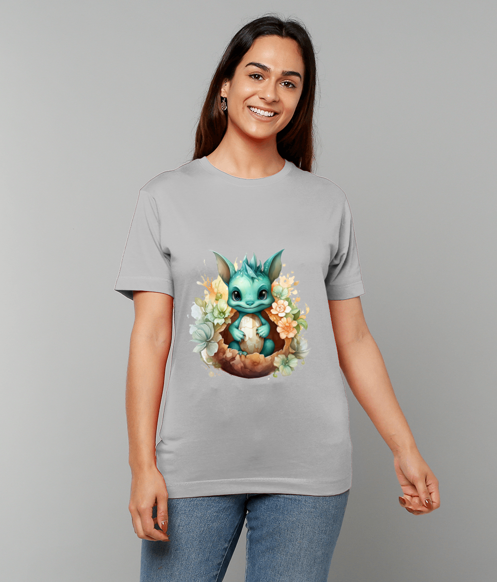 A turquoise baby dragon sits in a cracked eggshell surrounded by pastel flowers printed on a gray T shirt worn by a smiling woman standing against a light gray background.