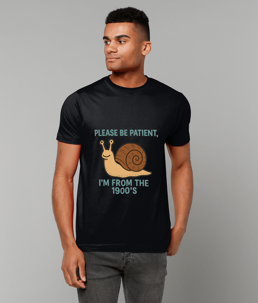 A young man wearing a black t-shirt that reads "PLEASE BE PATIENT," above a cartoon snail and "I'M FROM THE 1900'S", stands with hands relaxed, looking right against a plain gray background.