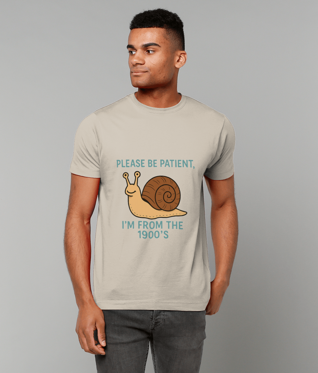 A man wears a light beige T-shirt depicting a cartoon snail and the text "PLEASE BE PATIENT, I'M FROM THE 1900'S", standing against a plain gray background.
