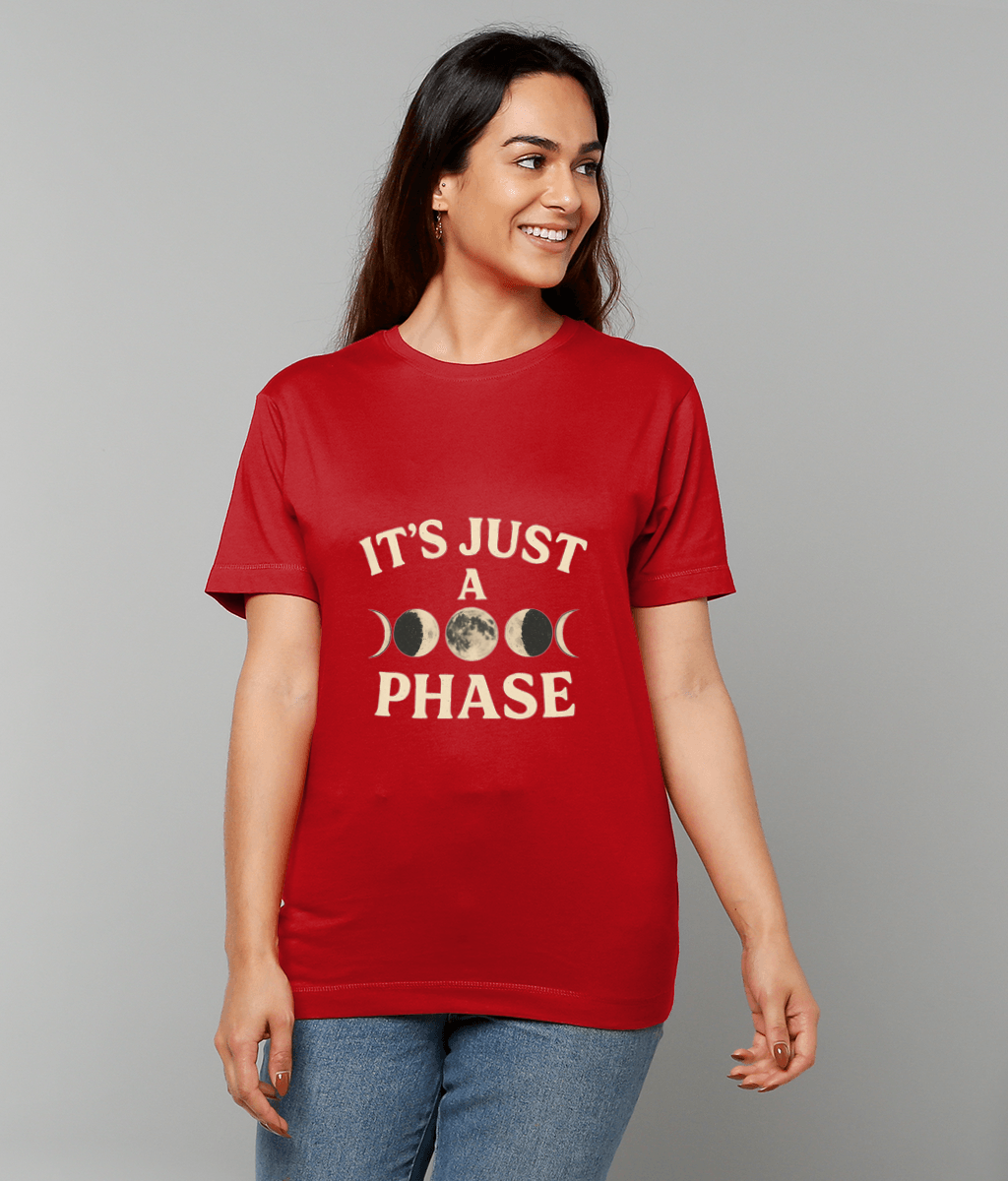 Red T-shirt reading "IT'S JUST A PHASE" with moon-phase graphics, worn by a smiling woman looking right, standing against a neutral gray backdrop and wearing blue jeans.