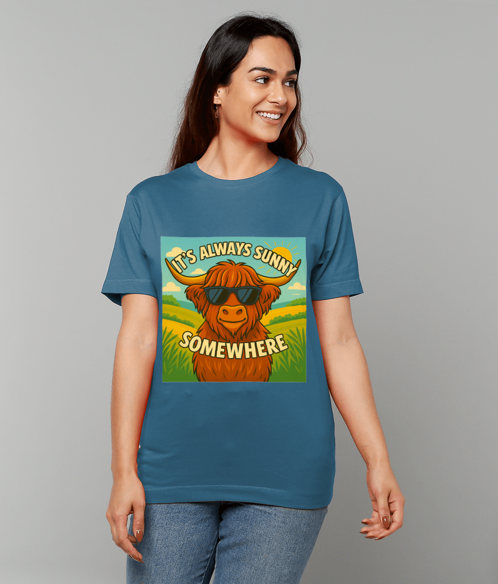 Woman wearing a blue T-shirt featuring a cartoon shaggy cow wearing sunglasses against rolling sunny fields with sun and clouds; text on the design reads "IT'S ALWAYS SUNNY SOMEWHERE".
