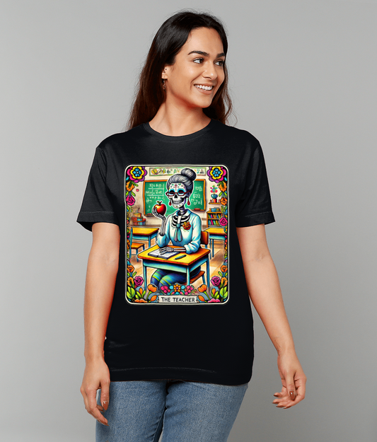 Person wearing a black T-shirt featuring a colorful Dia de los Muertos style skeleton teacher holding an apple at a classroom desk; text on design reads THE TEACHER.