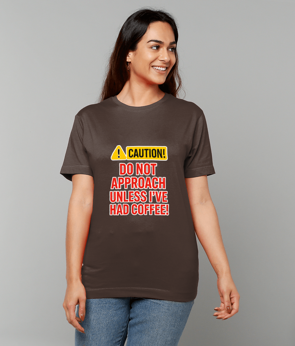 Person wearing a brown T-shirt smiling while standing against a plain gray background; shirt shows a yellow caution symbol and reads 'CAUTION! DO NOT APPROACH UNLESS I'VE HAD COFFEE!'