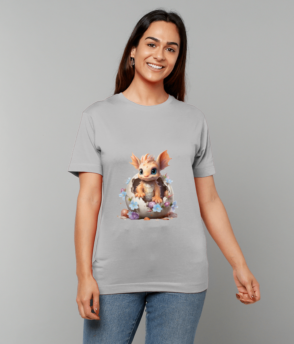 Baby dragon hatching from a cracked eggshell printed on a light gray T-shirt, worn by a smiling person standing against a plain gray backdrop.