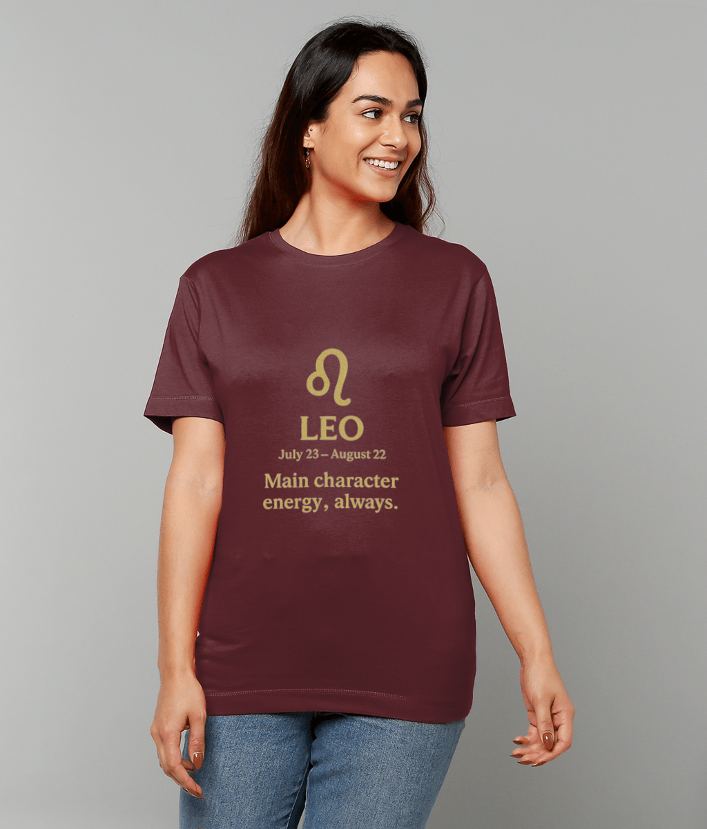Maroon T-shirt features printed text "LEO July 23 - August 22 Main character energy, always." worn by a smiling person looking right against a plain light-gray studio background.