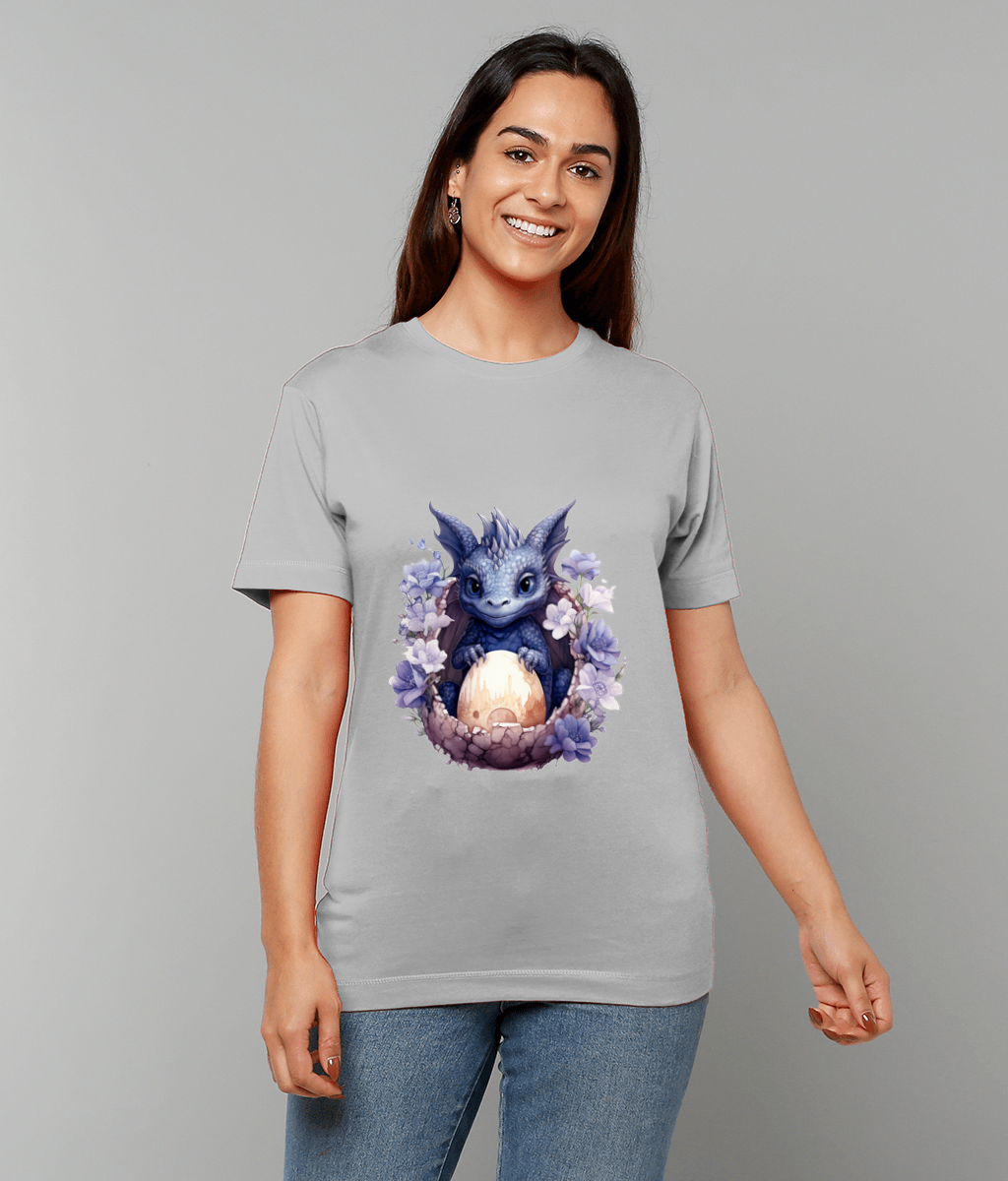 Small blue dragon cradles a hatching egg amid purple flowers on a light gray T-shirt worn by a smiling person against a plain gray background.