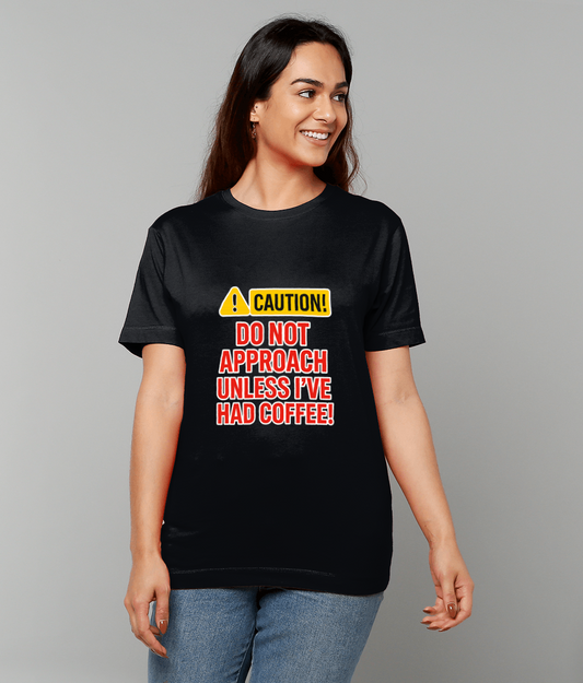 Black T-shirt displaying text "CAUTION! DO NOT APPROACH UNLESS I'VE HAD COFFEE!" worn by a smiling woman standing casually against a plain light-gray background.