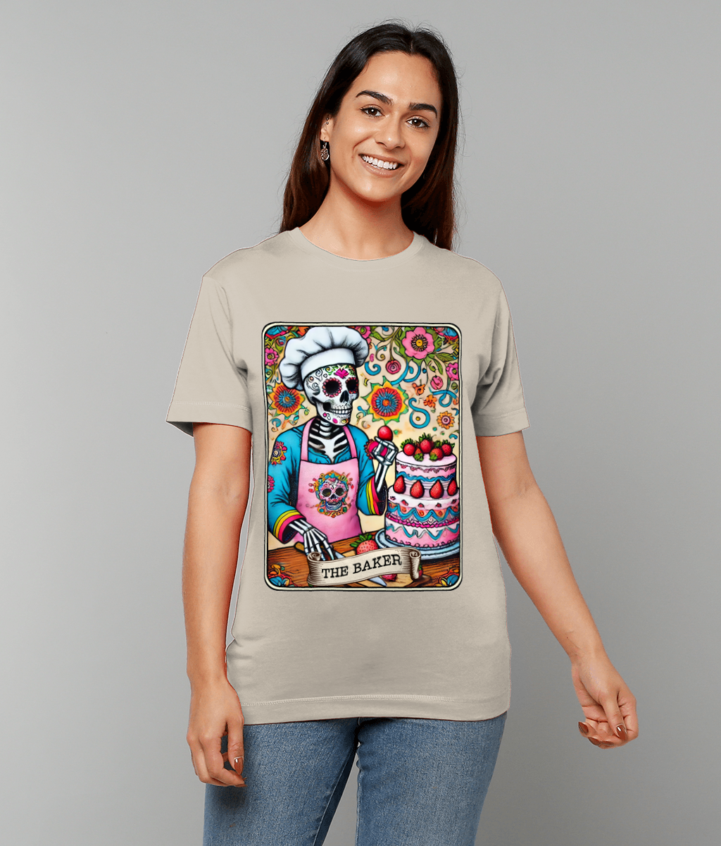 A skeleton baker is frosting a strawberry-topped cake against a colorful floral background printed on a beige T-shirt worn by a smiling woman, text on design reads THE BAKER.