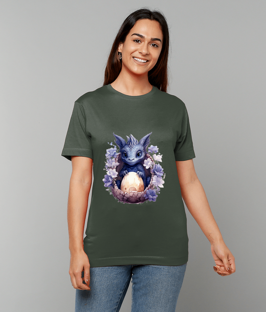 Smiling young woman stands against a plain gray backdrop wearing an olive green T-shirt featuring a blue baby dragon cradling a glowing egg surrounded by purple flowers.