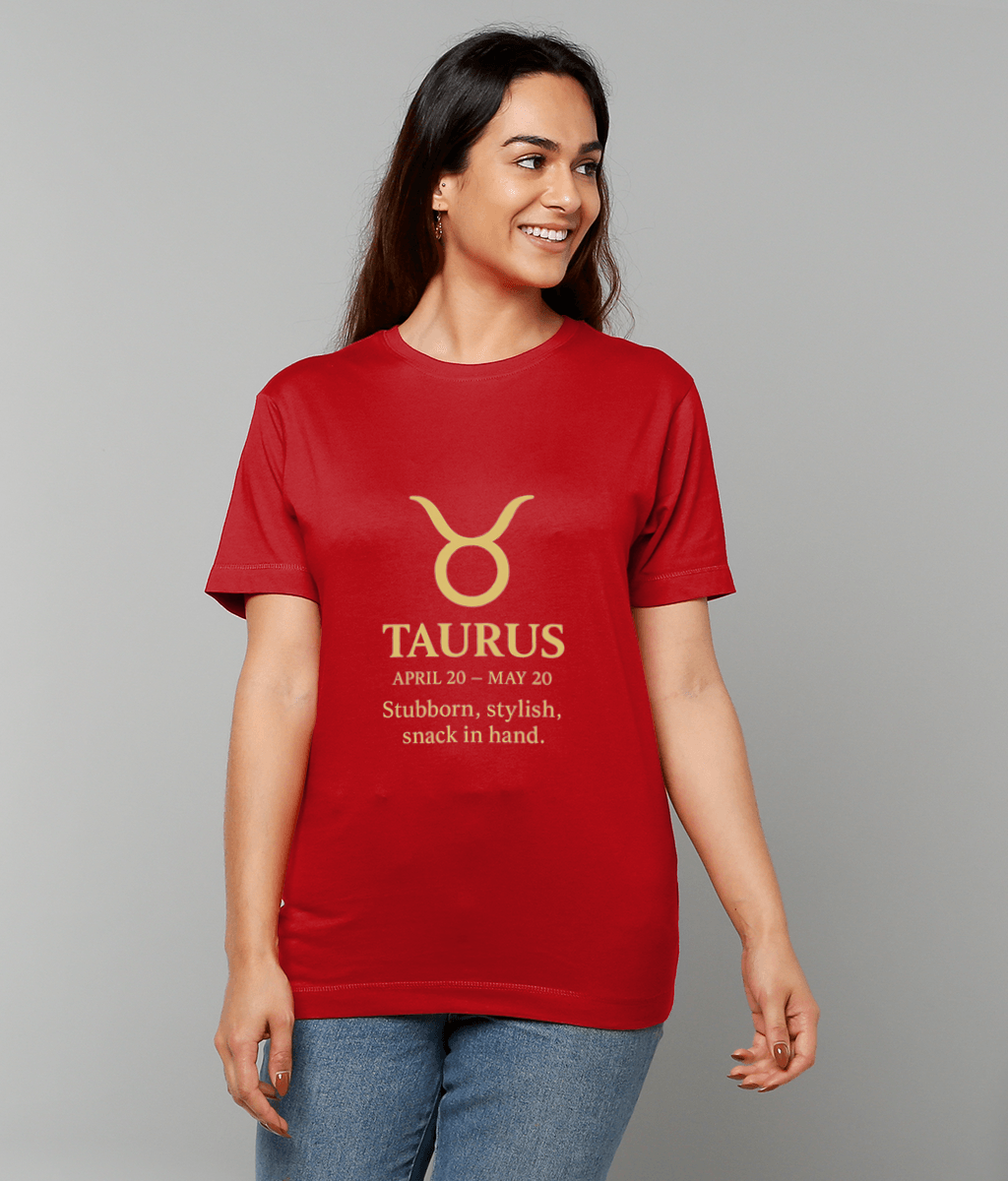 Woman wearing a red T-shirt featuring a gold Taurus symbol and the text "TAURUS APRIL 20 - MAY 20 Stubborn, stylish, snack in hand." smiles while standing against a plain gray studio background.