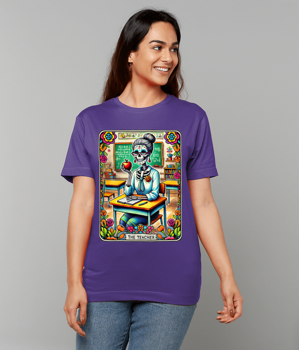 Day-of-the-Dead skeleton teacher illustration holding an apple while seated at a classroom desk printed on a purple T-shirt worn by a smiling person against a plain gray background Text: THE TEACHER