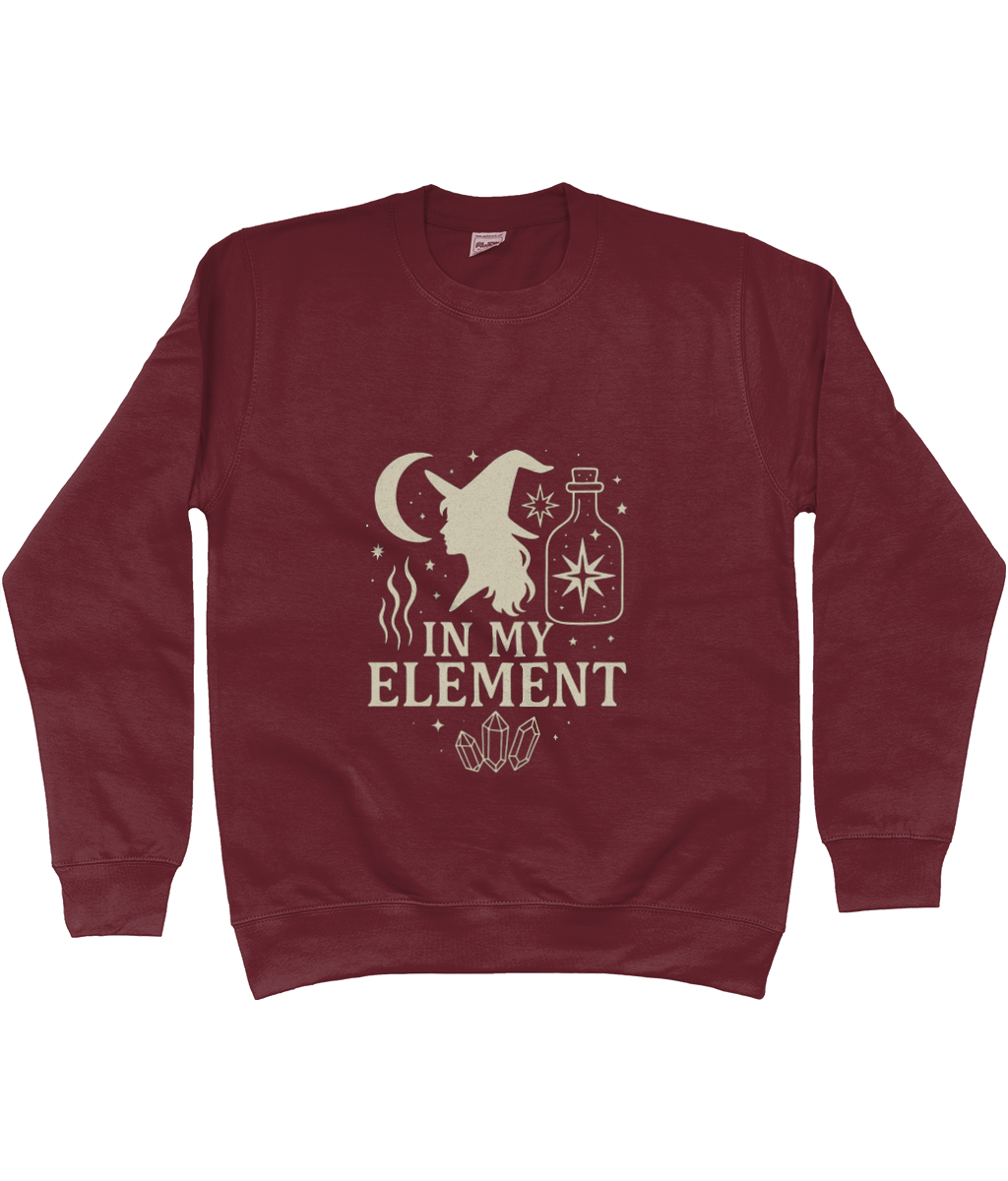 Maroon sweatshirt displays a cream witch silhouette wearing a pointed hat beside a crescent moon, potion bottle, crystals and sparkles with the printed words "IN MY ELEMENT", centered on a plain background.