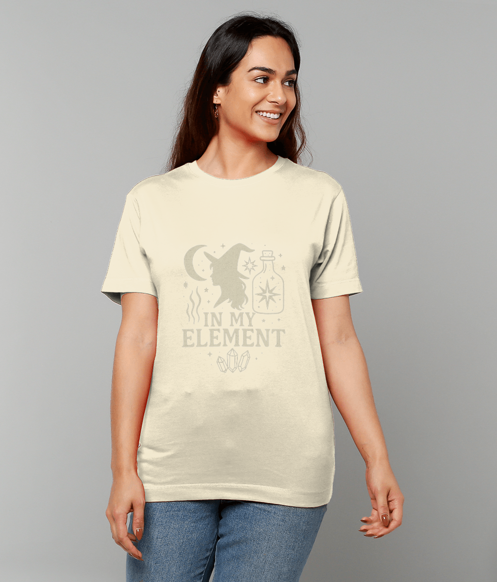 Young woman wearing a cream T-shirt displaying a witch silhouette, moon, potion bottle and crystals that reads IN MY ELEMENT, smiling and looking right in a neutral gray studio setting.