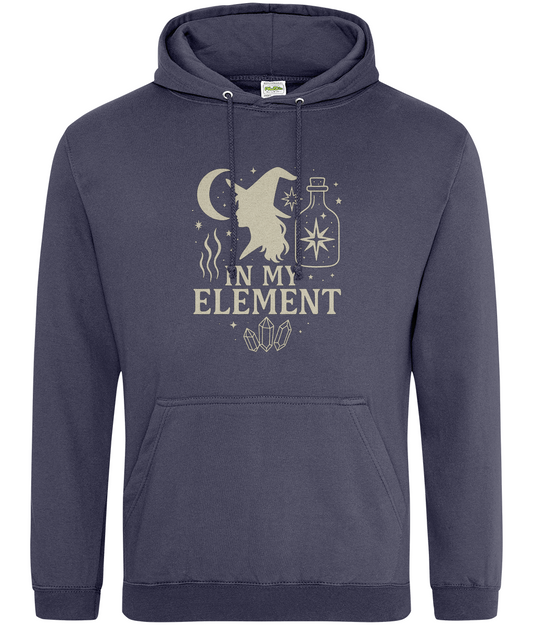 Navy hoodie displays a cream occult-style graphic of a witch silhouette, crescent moon, potion bottle, crystals and stars with the text IN MY ELEMENT centered on a plain white background.