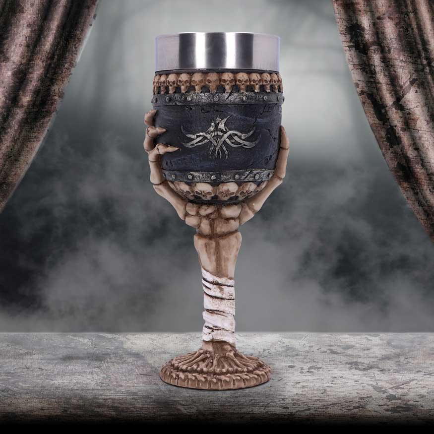 Bone chalice cradled by skeletal hands holds a metal-rimmed goblet, resting on a worn stone table amid low fog and framed by tattered curtains.