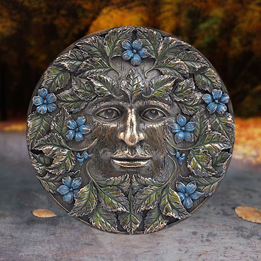 A round, ornate plaque depicts a face entwined with green leaves and blue flowers, set against a blurred, autumnal forest background.