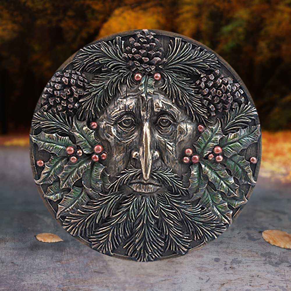 A circular, intricately carved coin features a face made of pinecones and foliage, with a solemn expression. It rests outdoors on a textured surface, against a backdrop of autumnal trees.