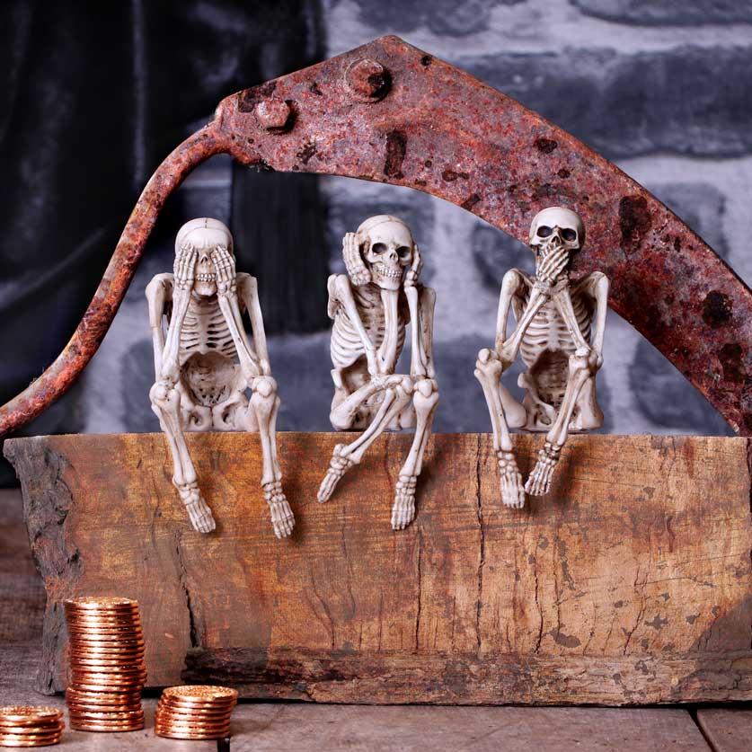 Three small skeletons sit atop a wooden block under a rusted metal piece, each posing "see no evil", "hear no evil", and "speak no evil". Coins are stacked on the block's side.