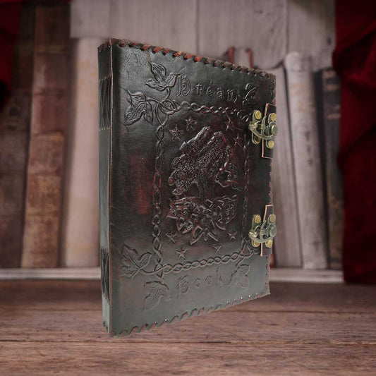 Leather-bound book with embossed leaf and wolf design, bearing text "Dream Book," features ornate brass clasps. Positioned standing on wooden surface, framed by blurred bookshelf backdrop.