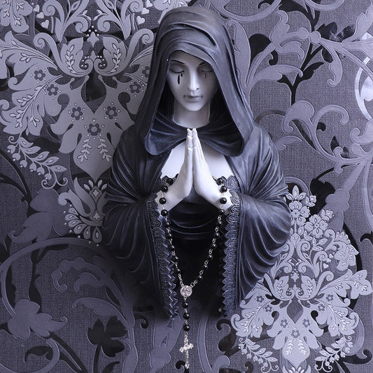 A statue of a hooded woman in prayer, adorned with a rosary, stands against an intricate floral-patterned background, creating a serene and contemplative atmosphere.