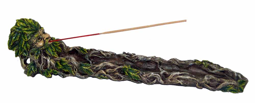 A decorative incense holder resembling a tree spirit face, adorned with green leaves. An incense stick is placed in the mouth, lying on a textured, wood-like base.