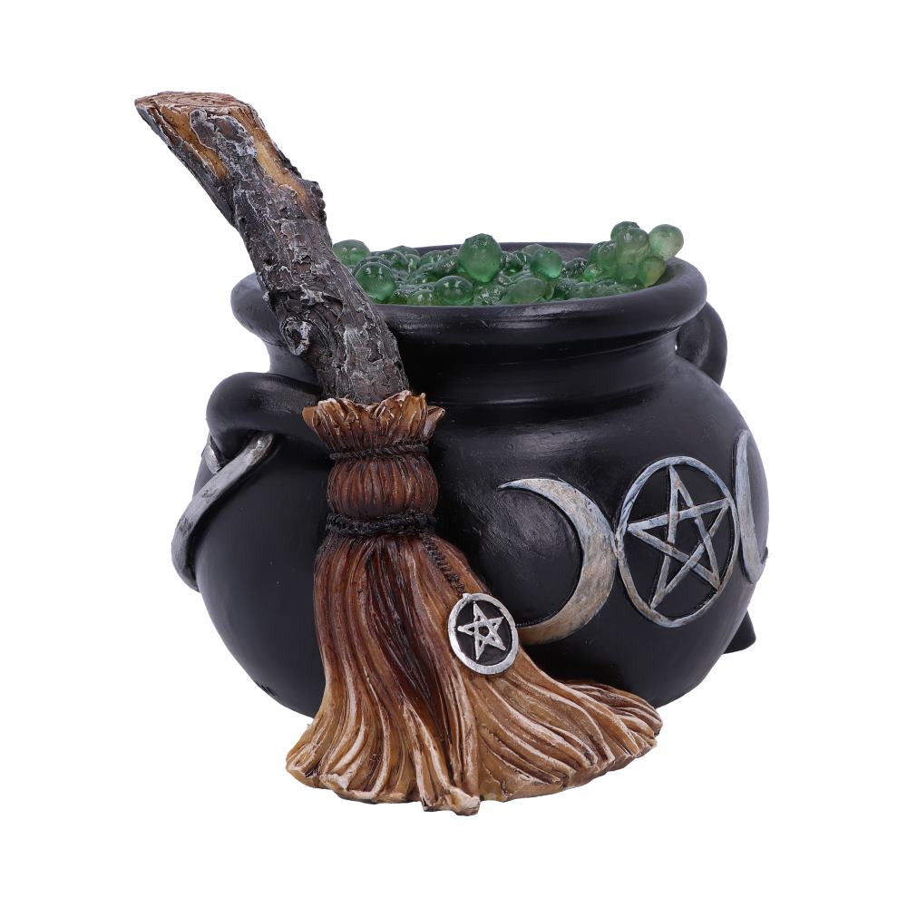 A black cauldron filled with green orbs sits with a wooden broomstick beside it; both adorned with crescent moons and pentacles, creating a mystical scene.