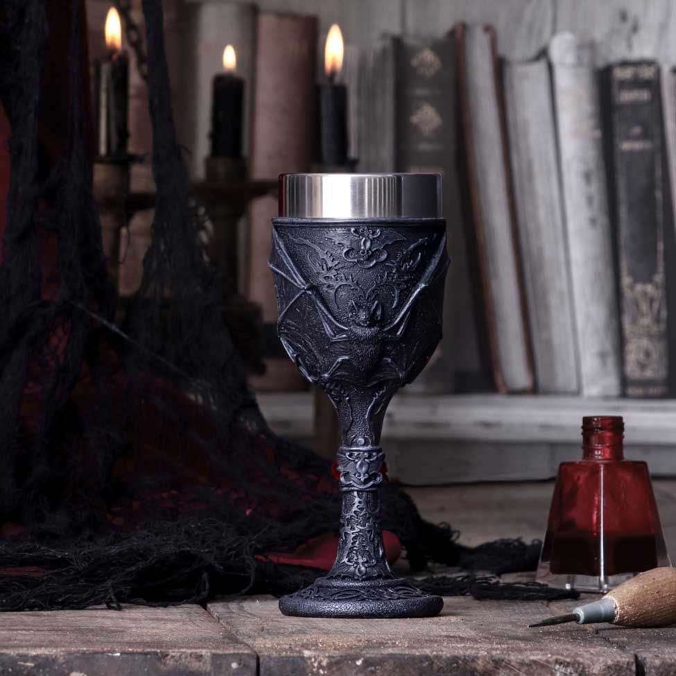 Ornate black goblet embossed with a bat stands on a wooden table amid lit candles, stacked antique books, a red ink bottle and carving tool beside dark draped fabric.