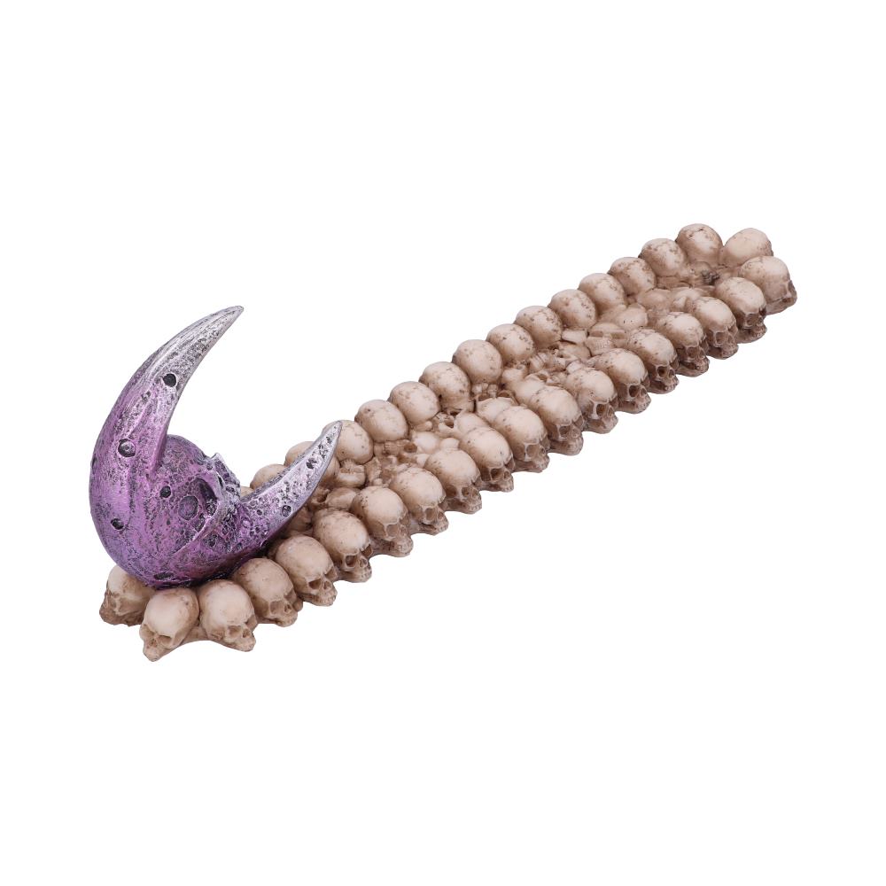 A purple crescent-shaped object rests on a linear arrangement of miniature skulls, creating a stark contrast. The skulls form a dense pathway against a plain, light background.