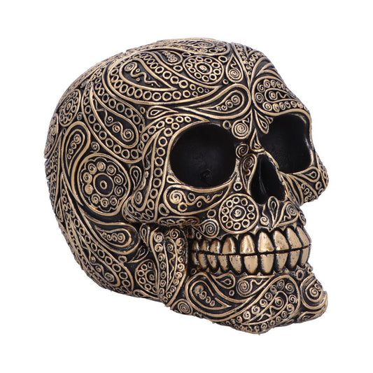 An intricately engraved metallic skull features swirling and circular designs, accentuating its eyes and teeth, set against a plain white background for emphasis.