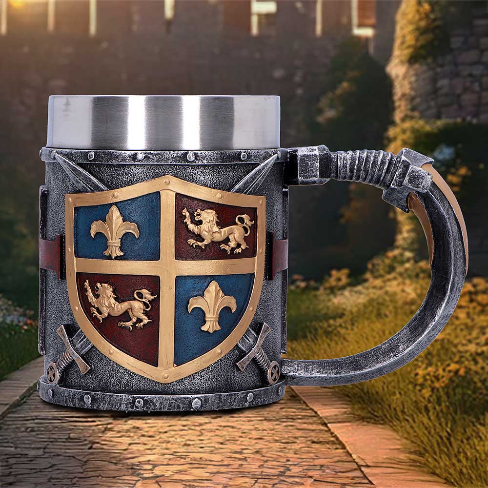A decorative medieval-style mug displays a gold-and-blue quartered shield with lions and fleurs-de-lis, sitting on a cobbled path before sunlit castle walls and grassy embankments.