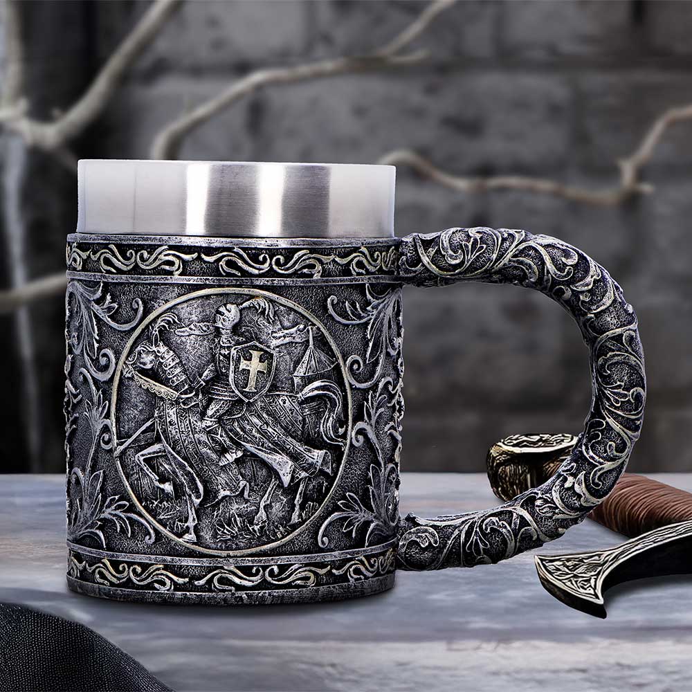 Ornate pewter tankard embossed with a knight on horseback medallion sits on a stone table beside a decorative dagger hilt against a blurred gray stone wall.