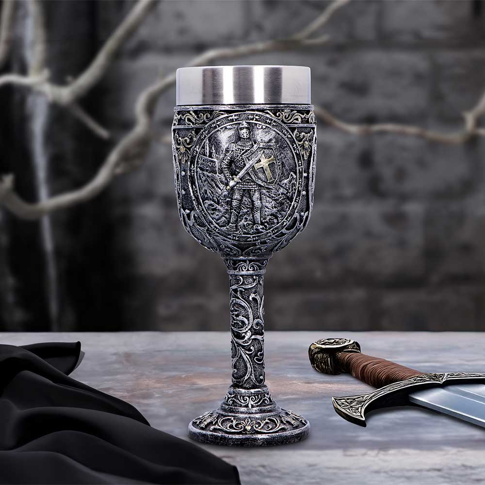 Ornate medieval goblet stands on a stone table, embossed with a knight; a decorative sword lies beside it and a folded black cloth rests left of a blurred wall.