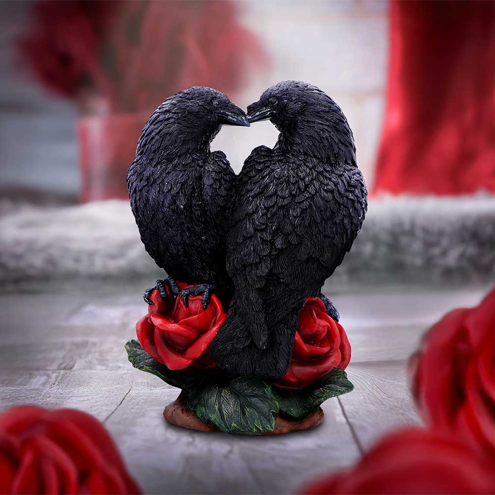 Two black raven figurines perched on red roses touch beaks forming a heart shape on a wooden tabletop with blurred red flowers in the background.