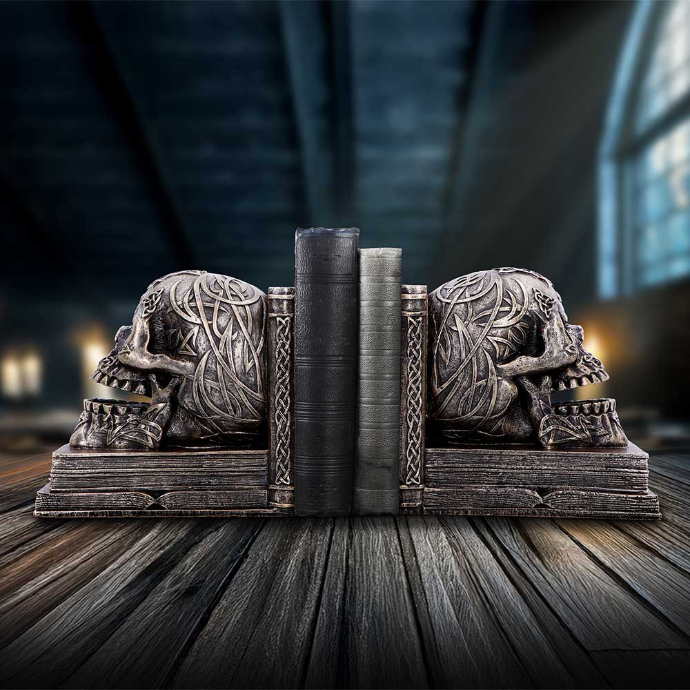 Carved skull bookends hold three leather-bound books upright on a wooden table in a dim vaulted room with blurred windows and warm lights.