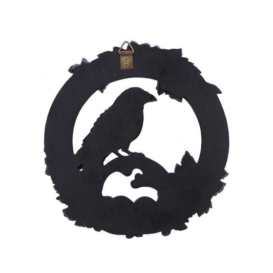 Black wooden circular plaque showing a perched crow silhouette cutout on a branch inside a leafy ring with a brass hanging loop, photographed on a white background.