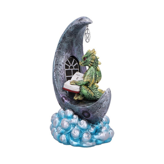 A small green dragon figurine reads an open book while seated inside a textured gray crescent moon perched atop pearlescent blue clouds, a window and dangling pentacle charm nearby.
