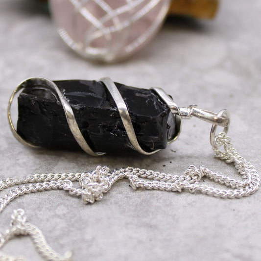 A black tourmaline crystal is wrapped in silver and attached to a silver chain, resting on a gray surface. In the blurred background are additional crystals.