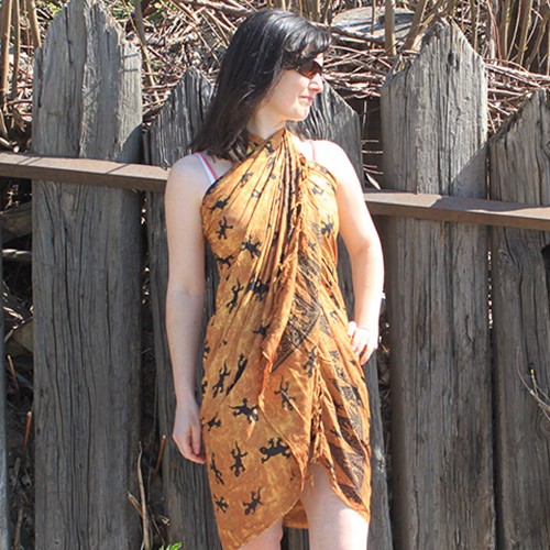 A woman is wearing a mustard-yellow sarong patterned with dark lizards, standing by a weathered wooden fence.