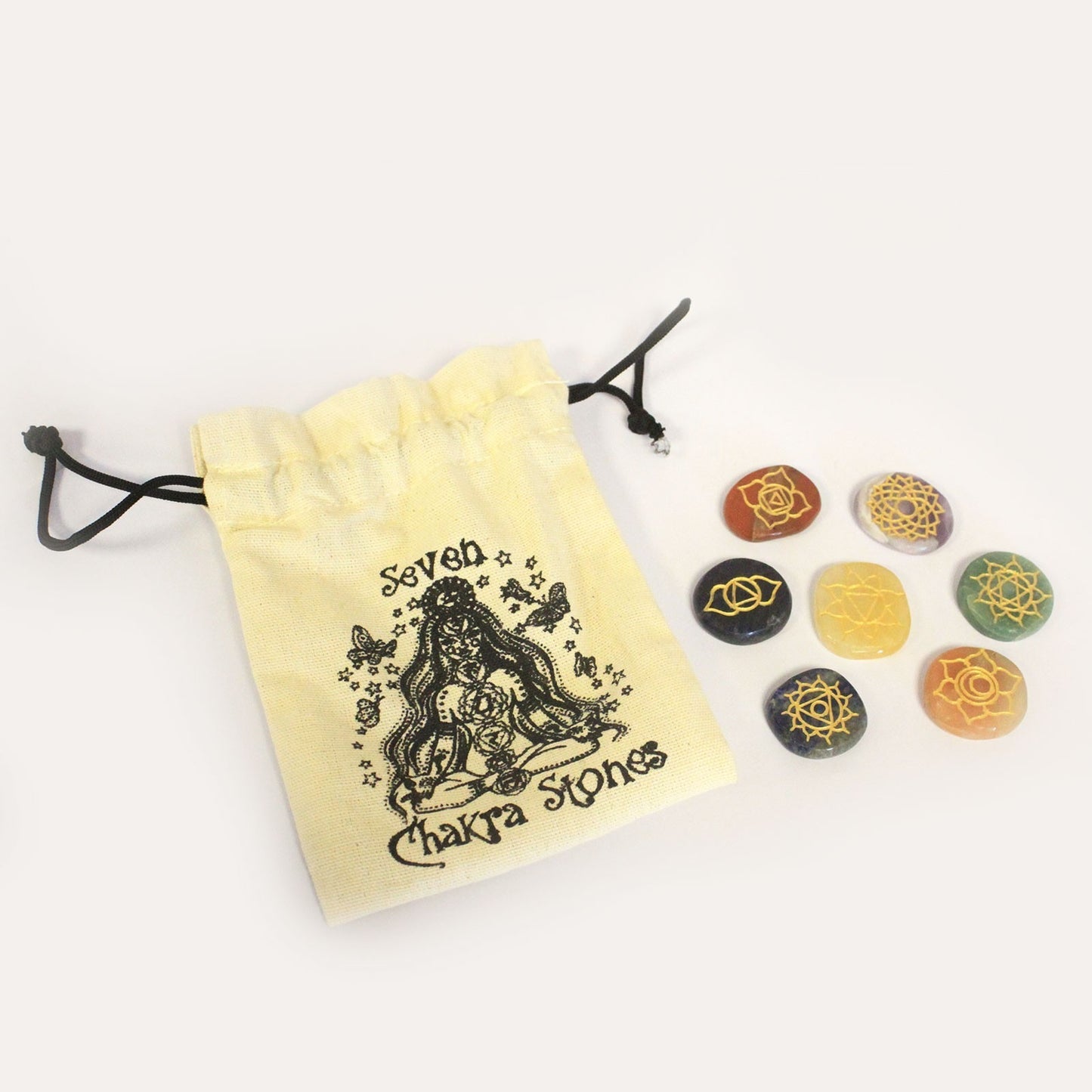 A small, pale yellow drawstring bag, printed with “Seven Chakra Stones” and an illustration, rests on a white surface beside seven colored stones, each displaying a different gold symbol.