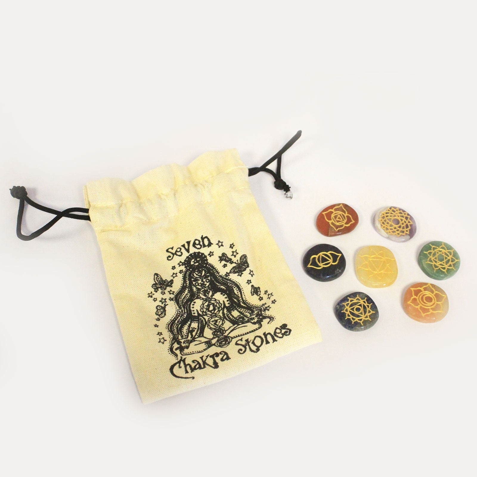 A small, pale yellow drawstring bag, printed with “Seven Chakra Stones” and an illustration, rests on a white surface beside seven colored stones, each displaying a different gold symbol.