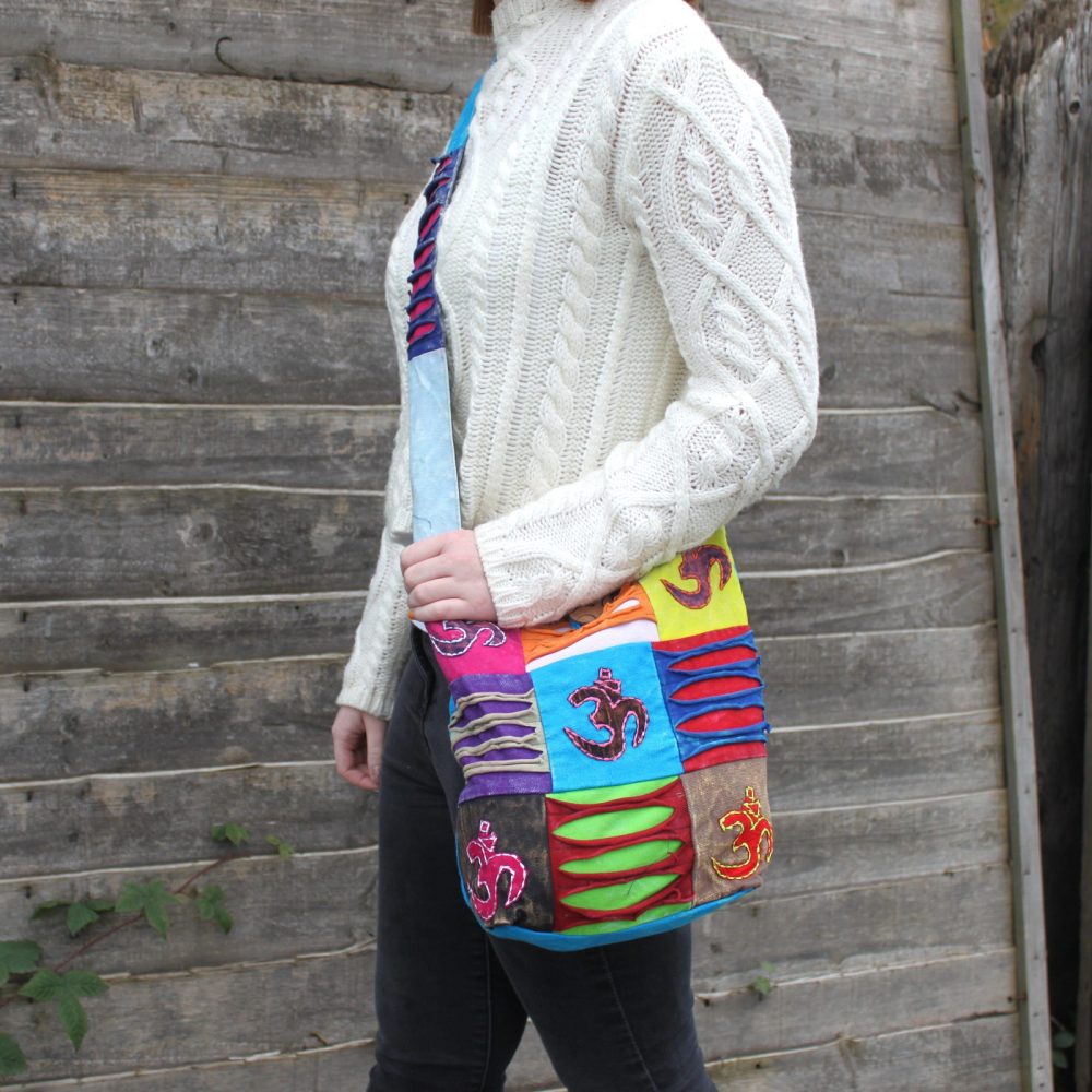 A patchwork shoulder bag with "Om" symbols hangs on a person wearing a cream-colored sweater and black pants. The person is standing near a wooden fence.