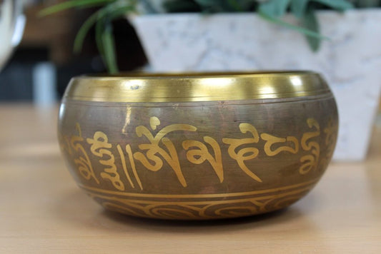 A brass singing bowl with engraved symbols sits on a wooden table. In the background, a marble planter with green leaves adds a natural touch to the scene.